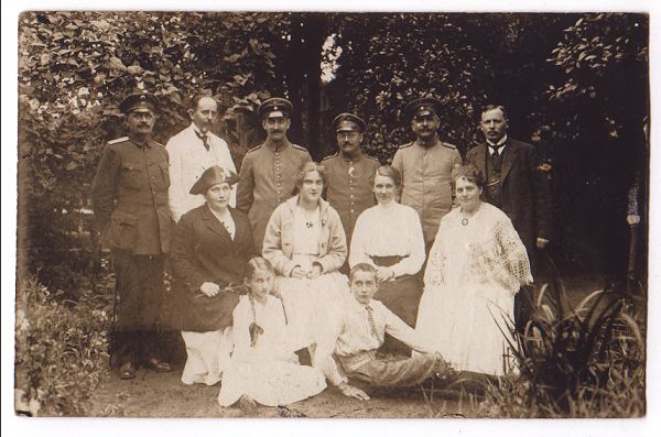 In a corner of the garden, a family poses for a group portrait.  At the back stand 6 men, 4 in uniform.  In front of them sit 4 women of varying ages, and at the front, a young boy and girl sit on the ground.