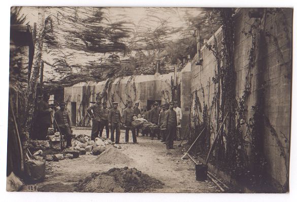 A view along a wide, curving, concrete-walled trench, roofed with camouflage netting.  A group of men stand in the middle distance, two of them carrying a stretcher bearing a sick or wounded soldier.